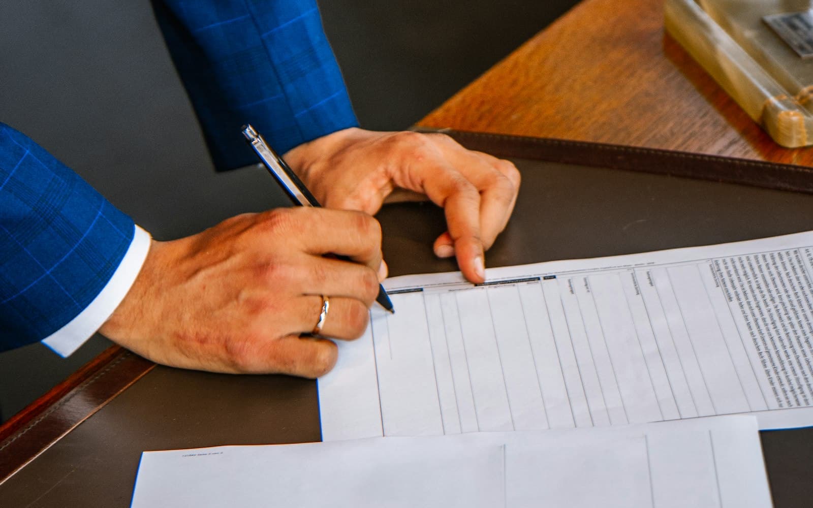 A person in a blue suit fills out a printed form with a pen at a desk