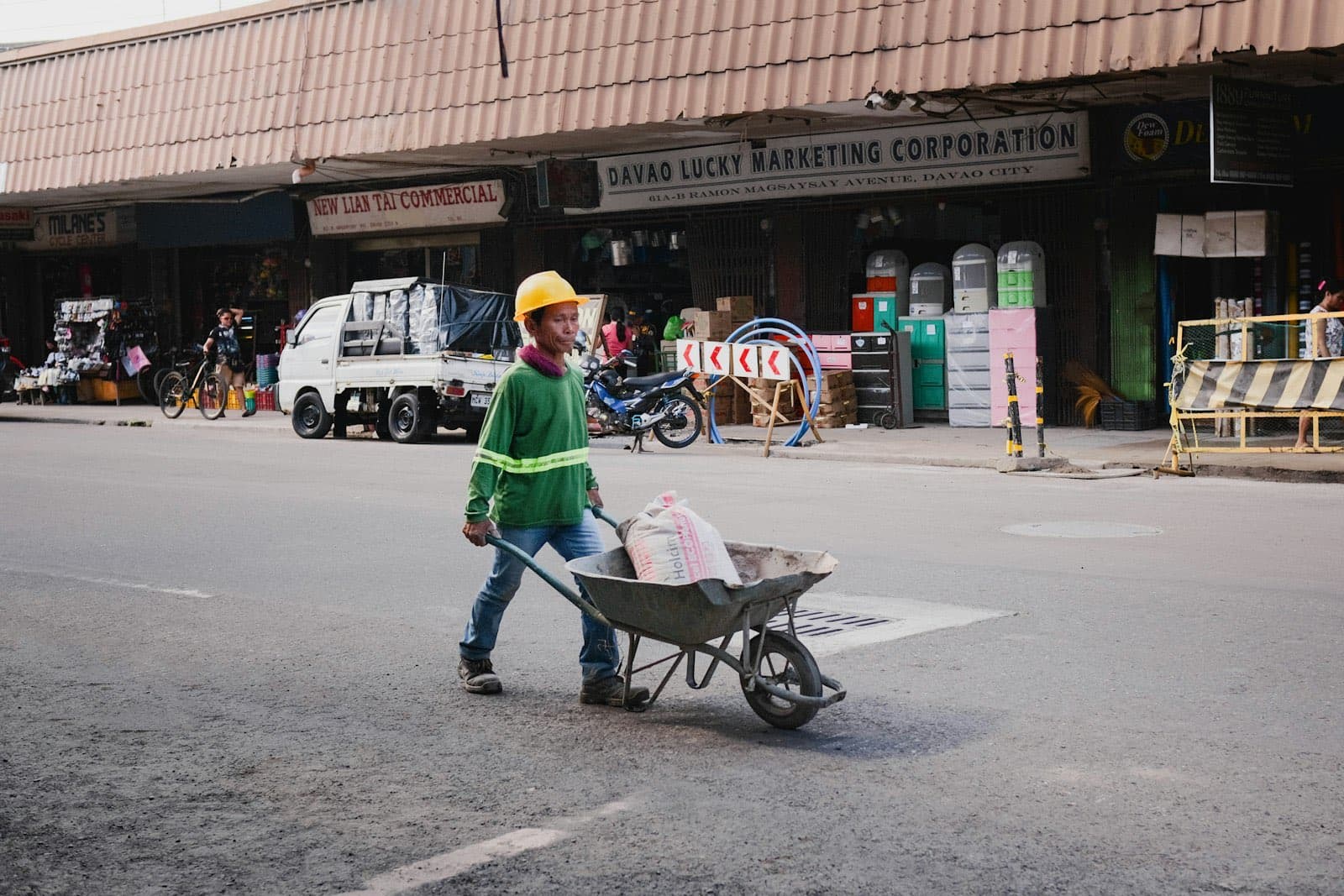 A construction worker in a hard hat and green vest pushes a wheelbarrow down a street in Davao City