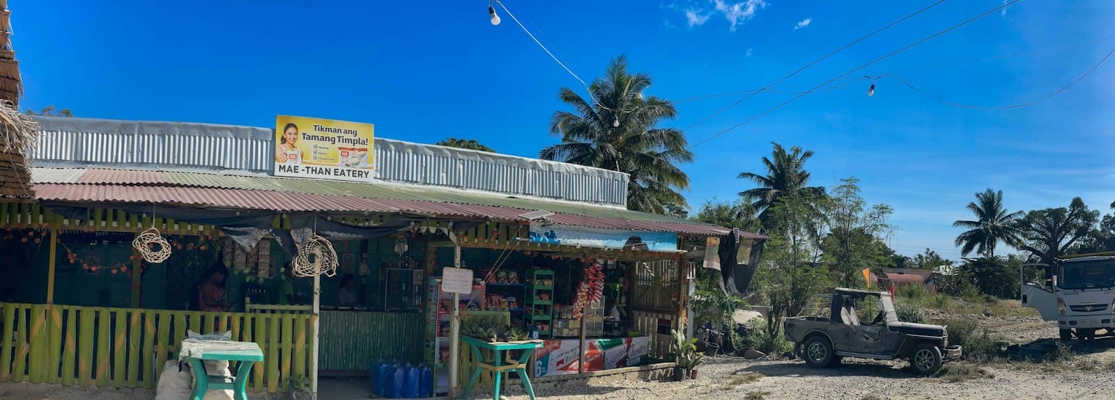 A roadside sari-sari store and eatery with a corrugated roof under palm trees in rural Palawan