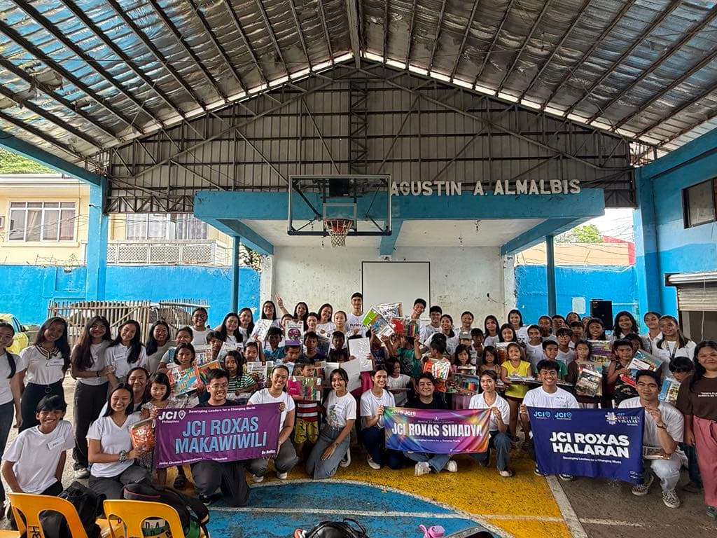 JCI volunteers pose with students and donated school supplies at a covered basketball court